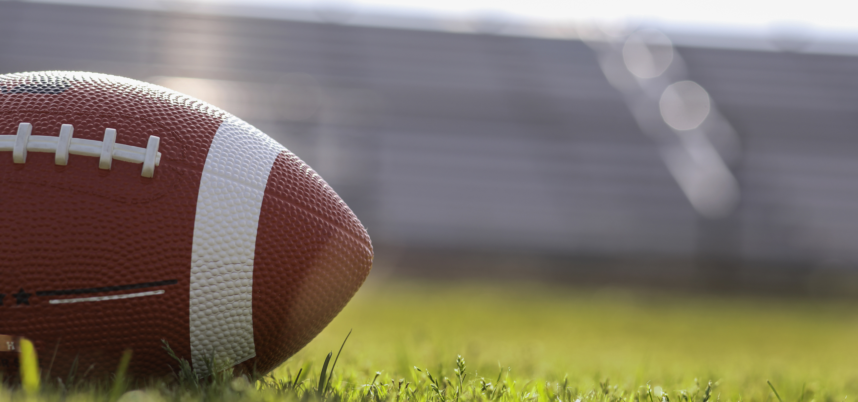 A close up shot of a football on the left, placed in the middle of a football field, with bleachers in the background and out of focus.